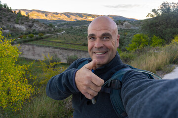 Hiker taking a selfie with thumbs up, outdoors.