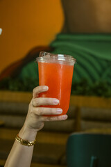 Close-up of a hand holding a cold red refreshing drink in a clear plastic takeaway cup.