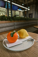 Close-up of two realistic fruit shaped gourmet desserts, one orange and one lemon, arranged on a ceramic plate in a modern cafe environment.