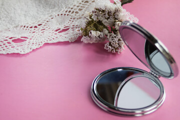 Compact mirror reflecting light on a pink background, featuring delicate dried flowers and white lace, symbolizing beauty, self care, and elegant vintage aesthetics