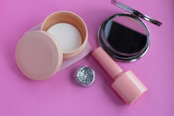 Beauty products and makeup accessories for self care routine, featuring pink nail polish, glitter, compact mirror, and face powder on a vibrant pink background