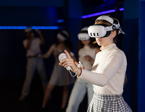 Young woman fully immersed in game using VR headset and controllers, standing in futuristic dark blue gaming room, actively gesturing to manipulate objects in virtual world