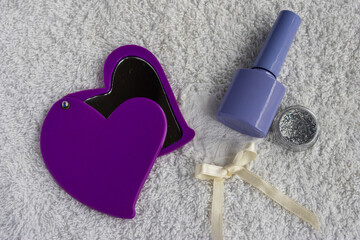 Purple heart compact mirror, nail polish bottle, and glitter on a soft white towel, symbolizing self care and beauty tools for manicure