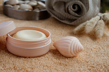 Self care essentials on a soft towel, featuring a creamy balm in an open container, a pink seashell shaped soap, and various spa accessories, promoting beauty and relaxation