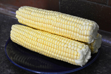 Raw corn on the cob on a dark plate. A delicious, ready-to-eat vegetable. A tasty and healthy dessert for both children and adults. A source of vitamins and fiber.