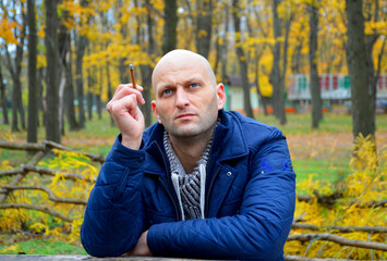 bald young man in a jacket smokes a cigarette on a bench in the autumn park. a man enjoys a calm rest, fragrant cigarette smoke and a good autumn day