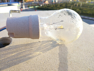 The light bulb hangs against a light concrete wall, illuminated by sunlight. Subtle shadows and traces of time are visible on the surface, creating an atmosphere of simplicity and everyday life.