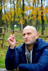 bald young man in a jacket smokes a cigarette on a bench in the autumn park. a man enjoys a calm rest, fragrant cigarette smoke and a good autumn day