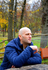 bald young man in a jacket smokes a cigarette on a bench in the autumn park. a man enjoys a calm rest, fragrant cigarette smoke and a good autumn day