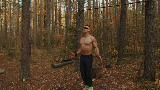 Young muscular man training in glade with horizontal bar nailed to trees in an autumn forest and performs jumping jack exercise. High intensity interval training Tabata. HIIT