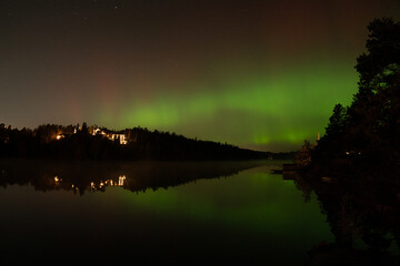 Green and red northern lights display over a lake near Stockholm, reflected in the water along with the surrounding forest and houses
