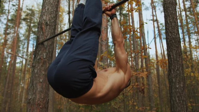 Young muscular man training on makeshift sports ground in autumn forest and performs leg raises on horizontal bar nailed to tree. High intensity interval training Tabata. HIIT