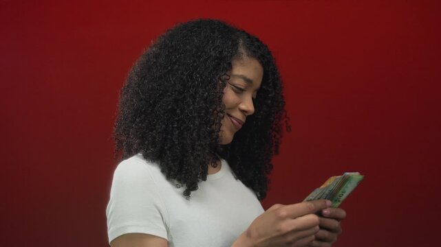 Woman smiling while counting rand banknotes and fanning cash stacks in vibrant red studio; confidence.