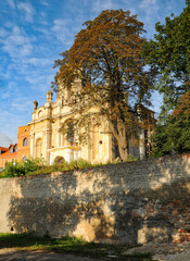 Fototapeta premium Historic Carmelite church in Bilshivtsi, Ukraine, with autumn foliage. The church sits atop a stone wall, bathed in golden sunlight against a blue sky with wispy clouds