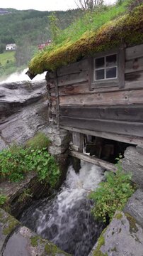Old restored wooden cabin with a green roof in the middle of the Hellesyltfossen waterfall in Hellesylt, Norway. Surrounded by colorful houses and lush forest