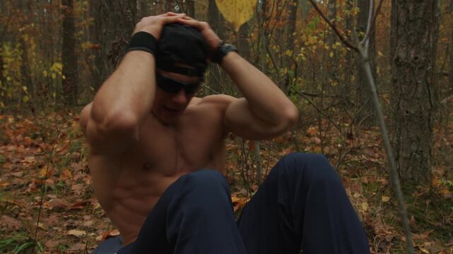 Shirtless bodybuilder training in an autumn forest and does crunches while lying down in glade covered with yellow leaves. High intensity interval training Tabata. HIIT