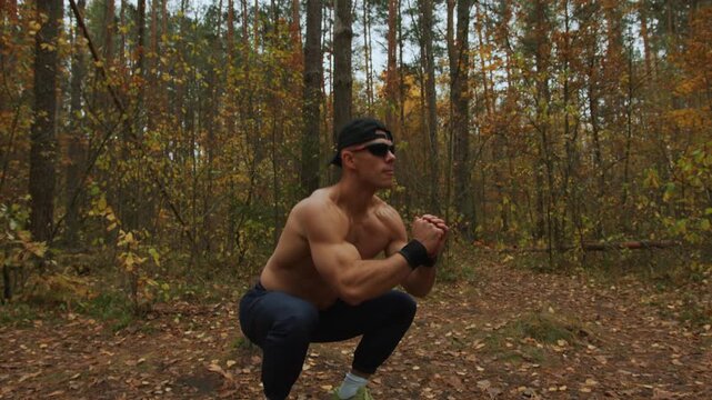 Young muscular shirtless man performs squats in an autumn forest clearing covered with yellow leaves. Calisthenics. Bodyweight exercises. Tracking shooting