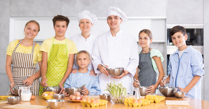 Successful experienced male and female chefs in white jackets and toques leading cooking class for interested teenage children, posing in workshop with group