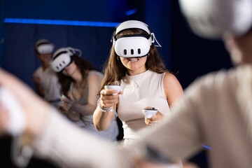 Cheerful emotional young female in VR goggles sitting in virtual reality room with group of friends, manipulating objects or shooting at targets with gaming controllers in hands.