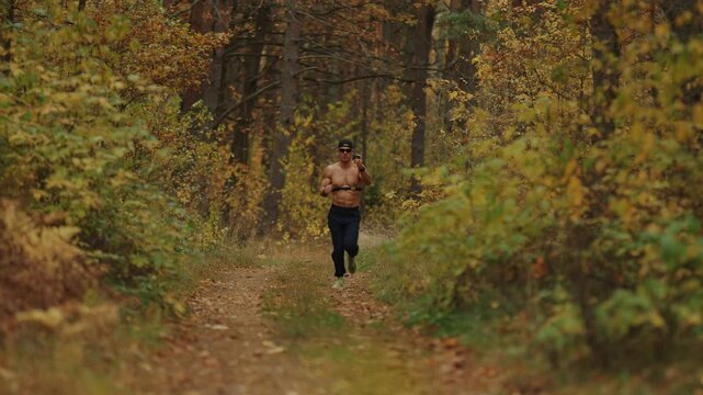 Young man wearing cap and sunglasses runs in an autumn forest with chest heart rate monitor and and watches indicators pulse by looking at monitor in his hand to control intensity of his workout