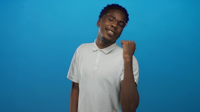 Man wearing white polo shirt and glasses casually points thumb backwards in studio against vibrant blue wall; confidence.