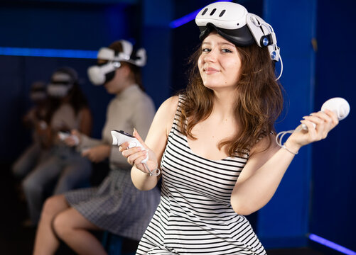Happy teenage girl with handheld controllers in hands and VR glasses on head sitting in virtual reality game room with blue futuristic lighting, ready for exciting experience with group of friends