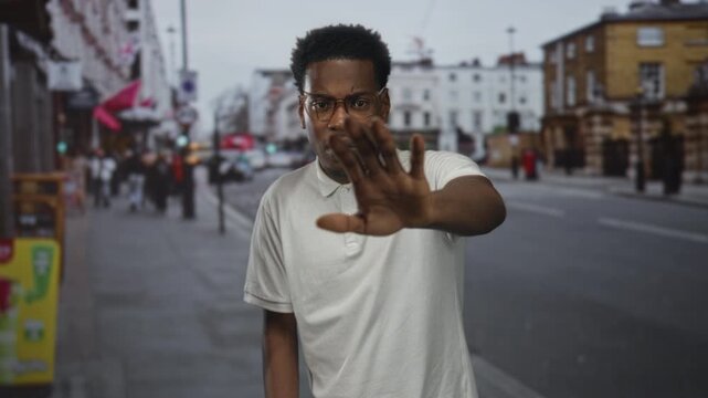 Man wearing glasses raises hand palm out toward camera on busy urban street crowded with pedestrians and cars, halting the view; defiance.