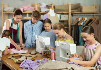 Smiling teen girl focusing on sewing on machine during hands-on lesson while classmates preparing patterns, fabric and fitting garments with female teacher
