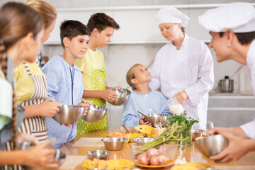 Interested tween boy taking part in cooking classes for children, mixing sauce in bowl with whisk, carefully listening to advice of professional chef instructor