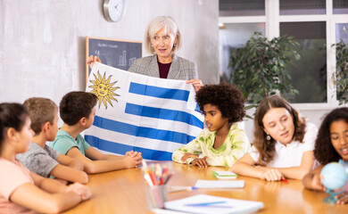 Mature female teacher, conducting a lesson in the classroom, tells the pupils the history of Uruguay and holds the national .flag of the country in her hands