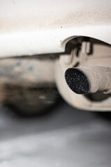 A close-up view of a car exhaust pipe releasing smoke in a city setting. The image captures the gray emissions against a blurred background, indicating pollution.