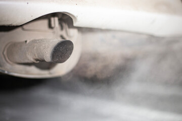 A close-up view of a car exhaust pipe releasing smoke in a city setting. The image captures the...