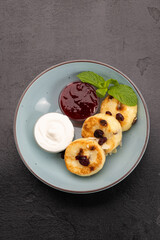Syrniki with sour cream and berry jam in a blue bowl on dark stone background