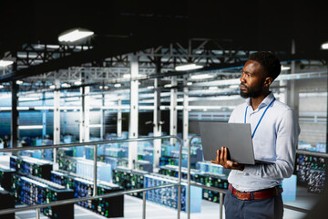 Server hub employee visualizing large language AI model using laptop, overseeing server room infrastructure from industrial platform. Data center technician reviews neural network.