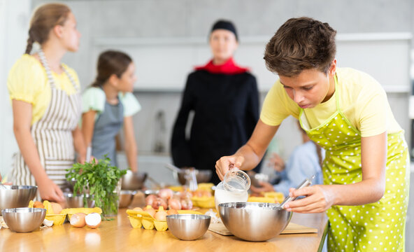 Interested enthusiastic teenager participating in culinary class of children group, engrossed in cooking process mirroring professional chef instructor steps