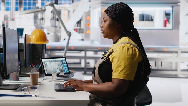 Technician monitors manufacturing data on pc in solar panel factory, employee finetunes equipment settings to support efficient solar power production. Woman seated next to assembly line. Camera B.