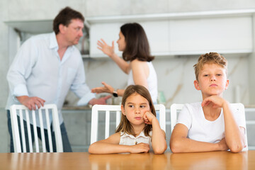 Children are sitting at kitchen table and hear their parents swearing, talking about divorce. Husband and wife are shouting at each other. Behind children are screaming parents in background, blurred