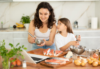 While cooking salmon fillet fish dish, woman and little daughter look at laptop screen and communicate with their grandmother via video link