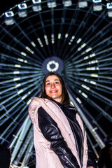 vertical portrait of a smiling young woman in front of a ferris wheel in an amusement park at night.