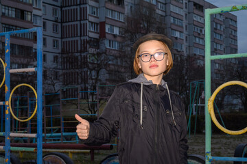 a boy in a cap and glasses shows a thumbs-up against the backdrop of an apartment building