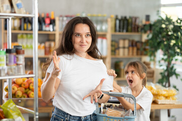 Exhausted and tired woman is outraged by her daughters behavior while shopping. Little girl pulls her mothers hand, demands to buy desired sweets