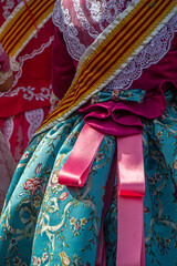 Close-up of a colourful traditional Fallas festival costume from Valencia with embroidered fabrics, pink satin ribbons, lace details, and a striped regional sash.