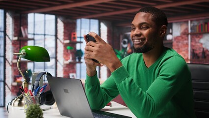 Man at home using smartphone to compose social media posts, producing content as a freelancer. African american guy monitoring profile engagement using mobile phone. Camera B.