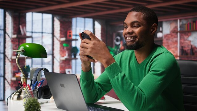 Man at home using smartphone to compose social media posts, producing content as a freelancer. African american guy monitoring profile engagement using mobile phone. Camera B.