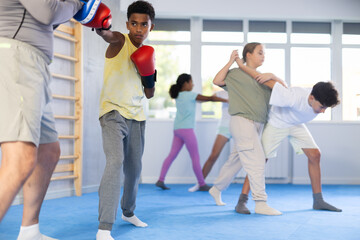 Motivated focused african american boy wearing boxing gloves working out with coach during group...