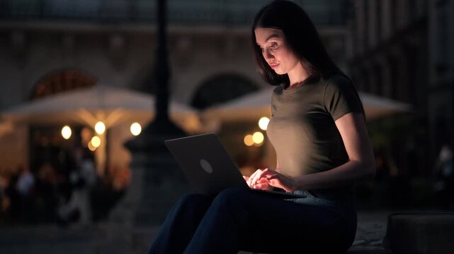 Young woman working on laptop outdoors at night, illuminated by screen light. Urban square with warm bokeh lights in background. Freelance, focus, modern lifestyle