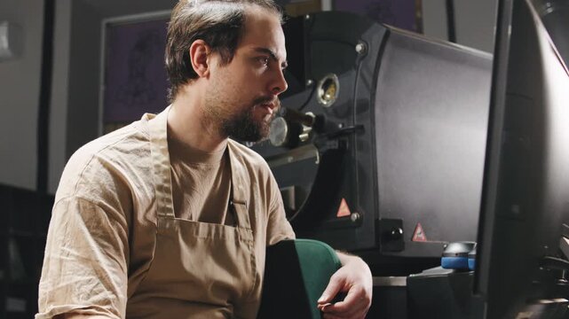 Coffee roaster working intently at a computer with machinery in a studio, focused on a project during the evening hours