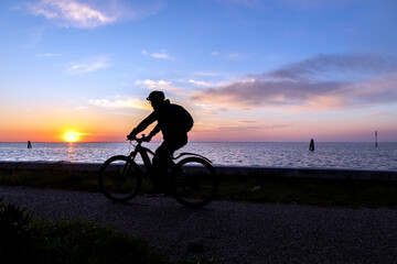 Silhouette of a cyclist riding along the waterfront at sunset on Pellestrina Island, Venetian Lagoon, Italy