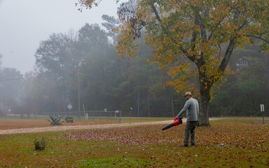 A person is seen blowing leaves in a yard on a foggy day. The autumn leaves are scattered on the ground, adding to the serene atmosphere.