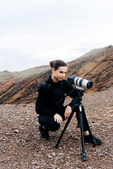 Female photographer in black attire crouches on rocky terrain, adjusting camera settings, capturing stunning landscape views, showcasing passion for travel photography and adventure